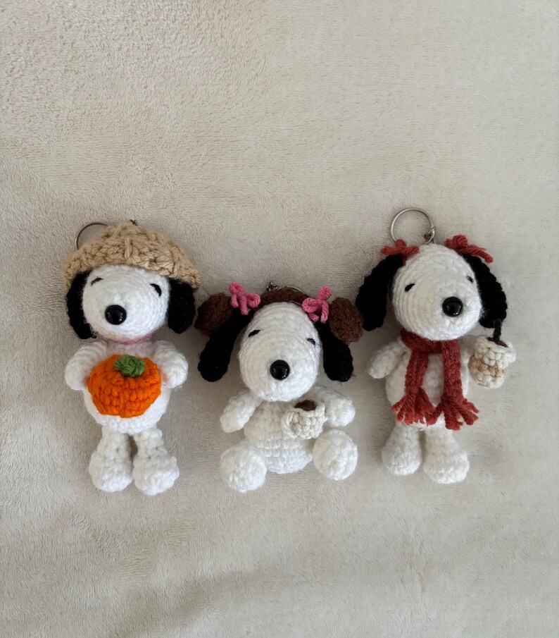 Op de afbeelding: Drie handgemaakte Snoopy-sleutelhangers van gehaakt garen. E&eacute;n sleutelhanger draagt een beige hoed en houdt een oranje pompoen vast. Een andere sleutelhanger heeft roze strikjes en een koffiekopje. De derde sleutelhanger heeft een rode sjaal en een koffiekopje.