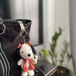 Op de afbeelding: Gehaakte Snoopy sleutelhanger met een rode sjaal en strik, met een koffiekopje. Het Snoopy figuur heeft zwarte oren en een zwarte neus. De sleutelhanger is bevestigd aan een zwarte tas.