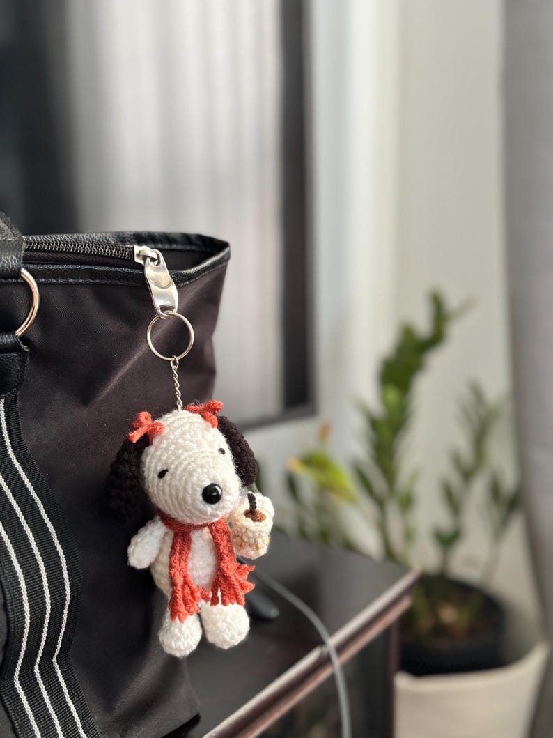 Op de afbeelding: Gehaakte Snoopy sleutelhanger met een rode sjaal en strik, met een koffiekopje. Het Snoopy figuur heeft zwarte oren en een zwarte neus. De sleutelhanger is bevestigd aan een zwarte tas.