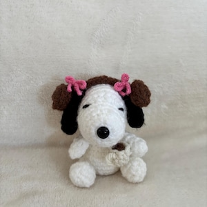 Op de afbeelding: Een handgemaakte gehaakte Snoopy-pop, met een witte body, bruine oren en zwarte accenten. De pop heeft roze strikken en houdt een kleine, beige beker vast. Het speelgoed staat op een zacht, wit oppervlak.