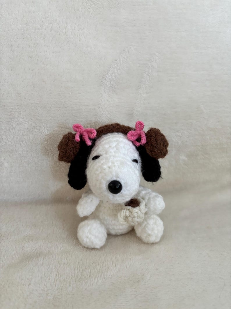 Op de afbeelding: Een handgemaakte gehaakte Snoopy-pop, met een witte body, bruine oren en zwarte accenten. De pop heeft roze strikken en houdt een kleine, beige beker vast. Het speelgoed staat op een zacht, wit oppervlak.
