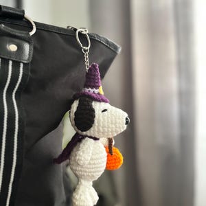 May include: Crocheted Snoopy keychain wearing a purple witch hat and scarf, holding an orange pumpkin. The white and black Snoopy is attached to a silver chain and is hanging from a black bag.