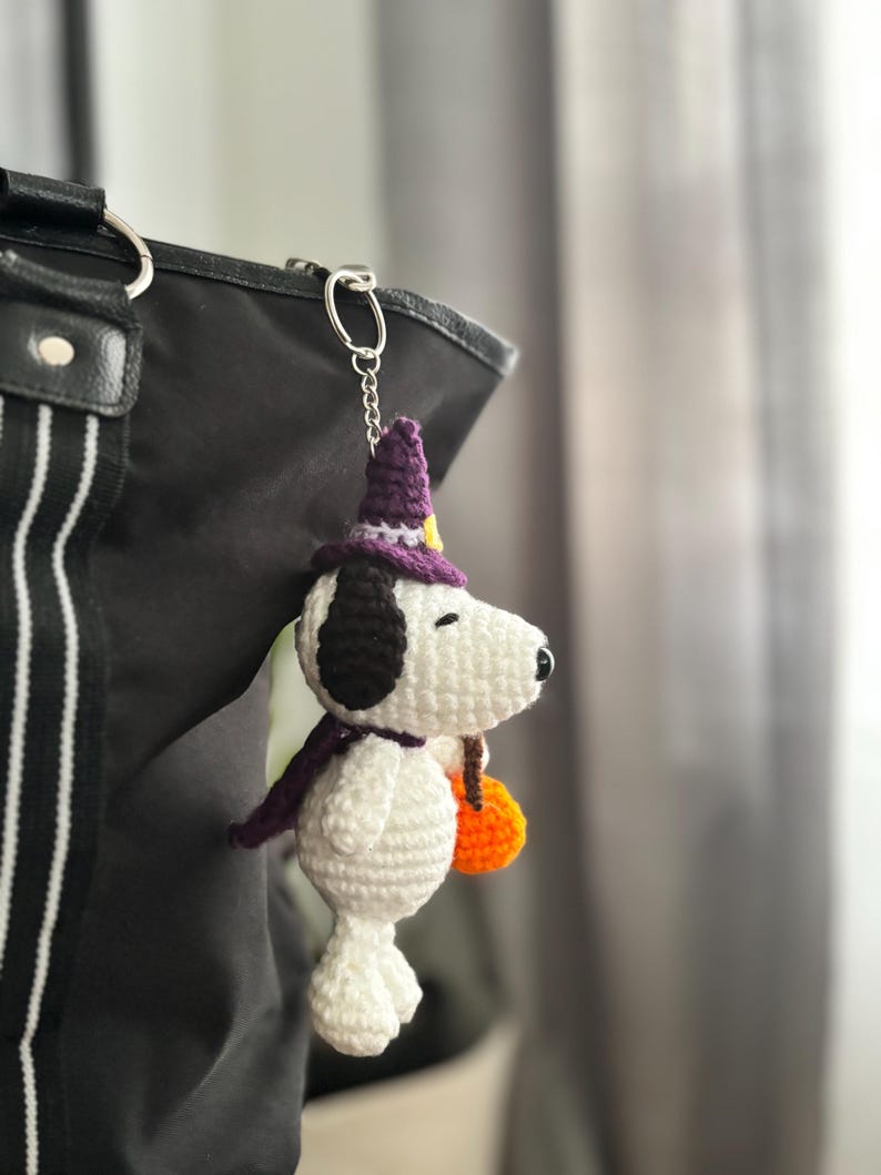 May include: Crocheted Snoopy keychain wearing a purple witch hat and scarf, holding an orange pumpkin. The white and black Snoopy is attached to a silver chain and is hanging from a black bag.