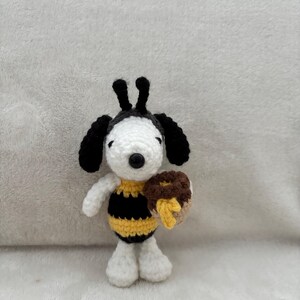 May include: A handmade crochet Snoopy doll dressed as a bee, with a black and yellow striped body and holding a brown donut with yellow honey. The doll has black ears and a black nose, and is standing on a white surface.