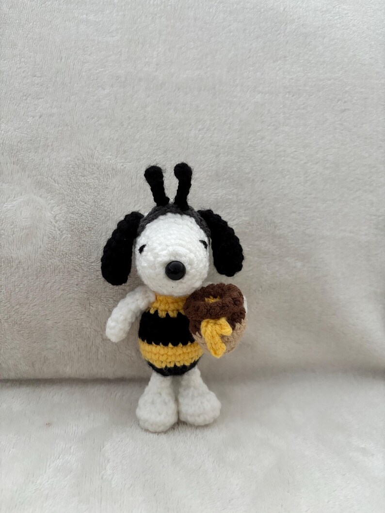 May include: A handmade crochet Snoopy doll dressed as a bee, with a black and yellow striped body and holding a brown donut with yellow honey. The doll has black ears and a black nose, and is standing on a white surface.