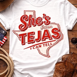 May include: White t-shirt with a red graphic that reads "She's from TEJAS I CAN TELL" in a Texas-shaped design. The shirt is surrounded by a cowboy hat, boots, rope, and an American flag.