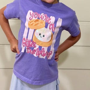 Puede incluir: Una camiseta morada con el texto "You're a Glitter Dumpling" en rosa y blanco. El gráfico presenta una caricatura de una bola de masa con una cara sonriente. La camiseta la lleva una persona con las manos en las caderas.