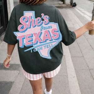 Puede incluir: Camiseta verde oscuro con un gráfico rosa y azul que dice "She's from Texas I can tell". La camiseta se combina con pantalones cortos a rayas rosas y blancas y zapatillas blancas. La persona camina por una acera.