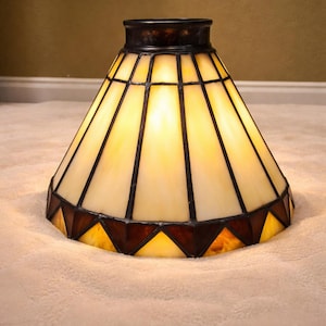 May include: A stained glass lampshade with a conical shape. The shade features a cream-colored background with dark brown geometric lines and accents. The bottom edge has a series of brown and amber-colored triangles.