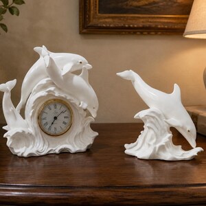 May include: A white porcelain clock featuring two dolphins leaping over a wave, accompanied by a separate white porcelain dolphin figurine emerging from a wave. The clock face has a gold rim, black Roman numerals, and black clock hands.