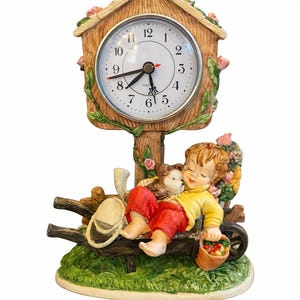 May include: A decorative clock featuring a child figurine resting in a wheelbarrow. The clock has a wooden-style frame with a round face and Roman numerals. The child is wearing a yellow shirt and red pants, holding a basket of fruit.