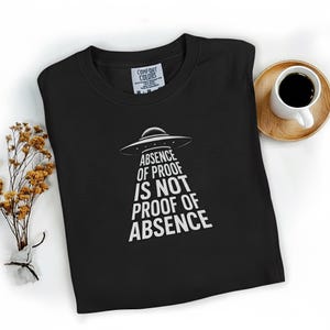 May include: Black crew neck t-shirt with a white graphic of a UFO and the text "ABSENCE OF PROOF IS NOT PROOF OF ABSENCE". The shirt is folded on a white surface, with a cup of coffee and a small bouquet of dried flowers.