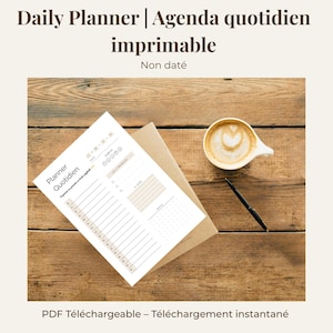 Peut inclure: Un planificateur quotidien imprimable avec le texte "Daily Planner | Agenda quotidien imprimable" et "Non daté". Le planificateur est sur une surface en bois avec une tasse de café et un stylo.