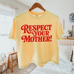 May include: A pale yellow short-sleeve t-shirt featuring the text "RESPECT YOUR MOTHER!" printed in a bold, red, distressed font across the chest. The garment has a crew neckline and is displayed on a wooden hanger against a neutral background.