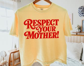 May include: A pale yellow short-sleeve t-shirt featuring the text "RESPECT YOUR MOTHER!" printed in a bold, red, distressed font across the chest. The garment has a crew neckline and is displayed on a wooden hanger against a neutral background.