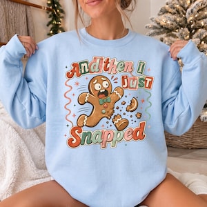 May include: A light blue crewneck sweatshirt featuring a graphic of a brown gingerbread person with a teal bow tie. The gingerbread person appears to be snapping in half. The text reads, "And then i just Snapped" in colorful, retro-style lettering.