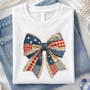 May include: White t-shirt featuring a patriotic bow design. The bow is made of patchwork denim, red gingham, and star-studded blue fabric. The shirt is folded on top of a pair of blue jeans.