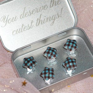 May include: An open silver tin with the text "You deserve the cutest things!" contains five star-shaped objects. Each star has a brown and blue plaid pattern. The tin sits on a pink, sparkly surface.