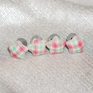 May include: Four star-shaped earrings with a plaid pattern in pink, white, and light blue. The earrings have gray posts. The earrings are displayed on a textured, silver surface.