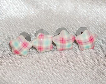 May include: Four star-shaped earrings with a plaid pattern in pink, white, and light blue. The earrings have gray posts. The earrings are displayed on a textured, silver surface.