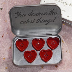 May include: Open silver tin with the text "You deserve the cutest things!" and five red heart-shaped buttons inside. Each button has four small holes for sewing. The tin is on a pink surface.