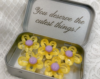 May include: A rectangular silver metal tin containing five yellow flower-shaped accessories with purple centers. The inside of the lid features the engraved text, You deserve the cutest things! The accessories have translucent yellow petals and smooth purple centers.
