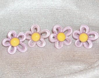 May include: Four daisy-shaped hair clips. Each clip features a light purple, open-petal design with a yellow center. The clips are arranged in a row against a shimmering, silver background.