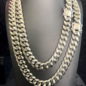 May include: A thick, polished silver-tone curb chain necklace featuring interlocking rounded links and a secure box clasp closure. The heavy metal chain displays a high-shine reflective finish, creating a bold, uniform appearance across the entire length of the jewelry piece.