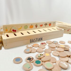 Könnte beinhalten: Holz-Lernspielzeug mit dem Namen "BASTIAN" auf der Seite. Das Spielzeug verfügt über Schlitze und Holzscheiben mit farbenfrohen Illustrationen von Früchten, Gemüse und Formen. Ein unterhaltsames und ansprechendes Lernwerkzeug.