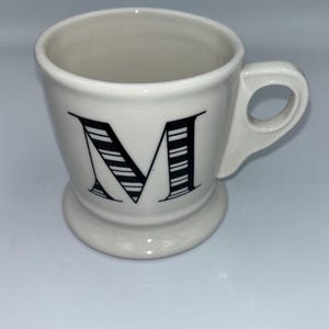 Op de afbeelding: Een witte keramische mok met een zwarte letter "M" op de voorkant. De letter is ontworpen met zwart-witte strepen. De mok heeft een handvat en een voet.