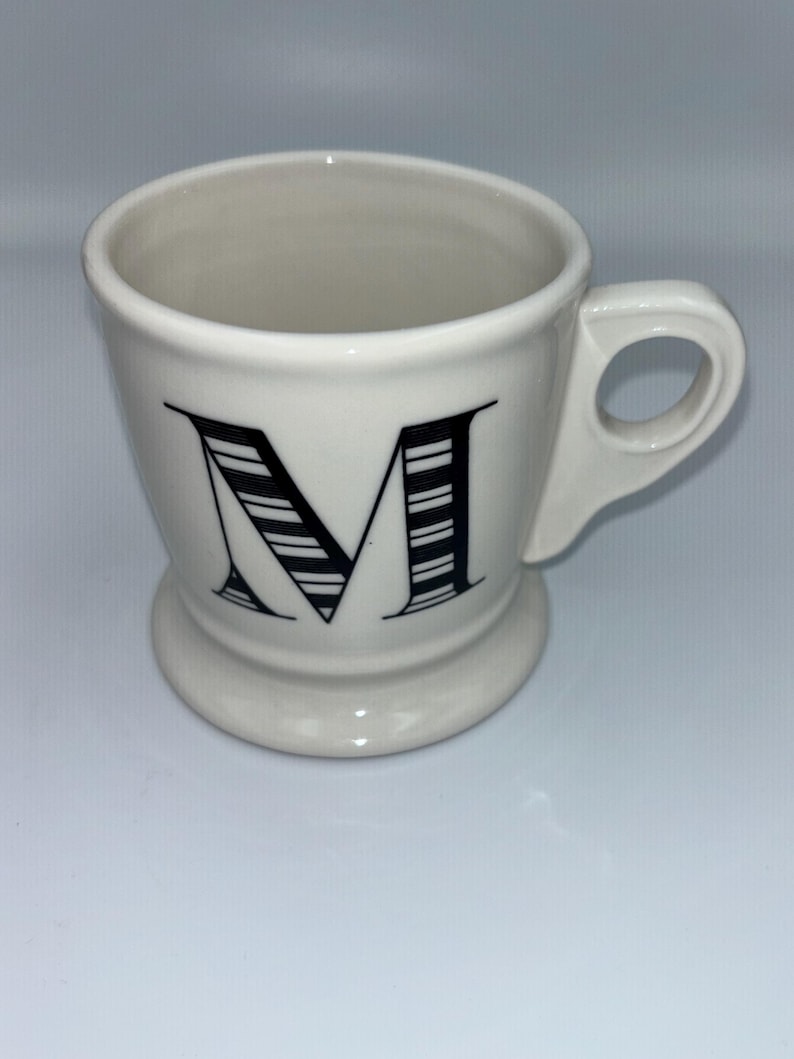 Op de afbeelding: Een witte keramische mok met een zwarte letter "M" op de voorkant. De letter is ontworpen met zwart-witte strepen. De mok heeft een handvat en een voet.