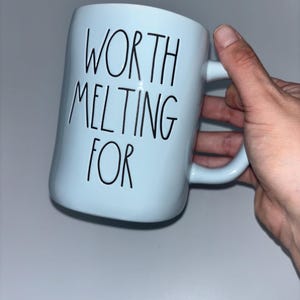 May include: A light blue ceramic mug featuring the black text "WORTH MELTING FOR" printed in a tall, thin, handwritten-style font. The mug has a smooth, cylindrical body and a curved handle on the right side, designed for holding hot beverages.