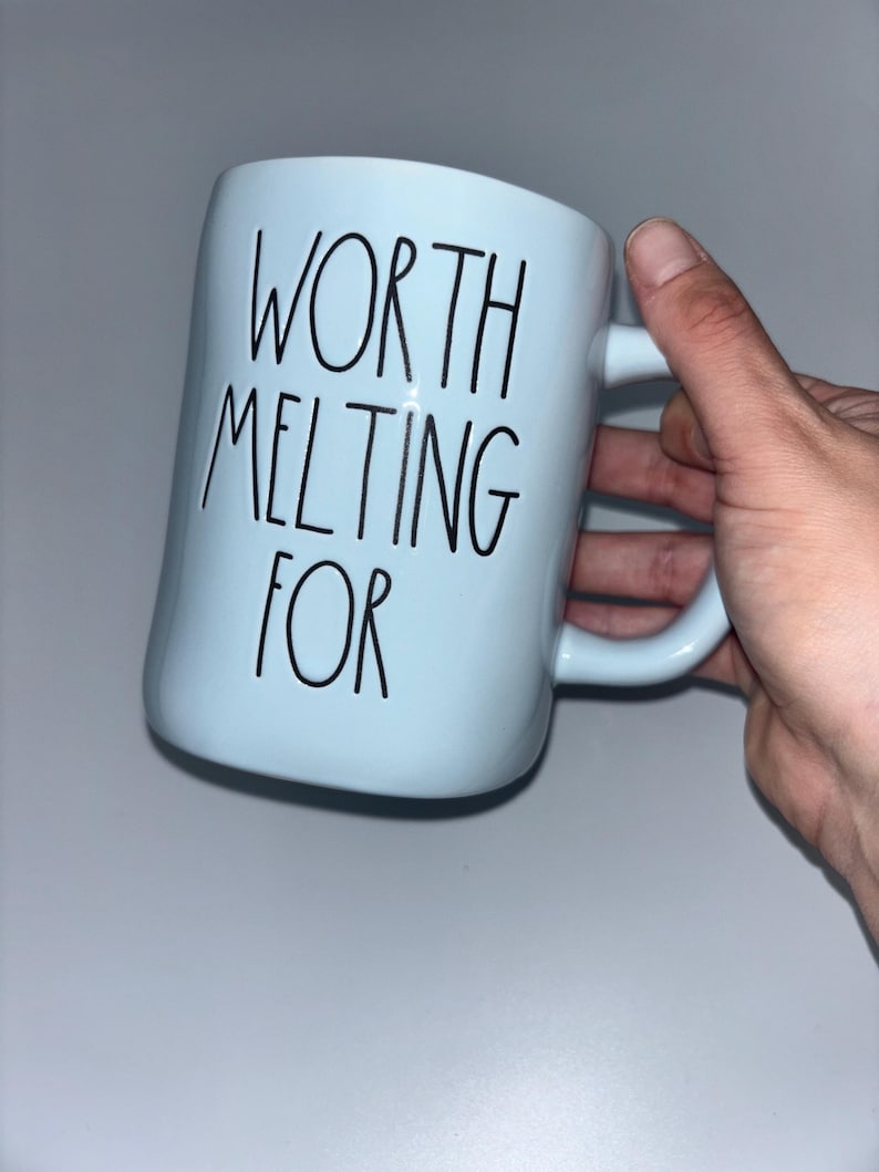 May include: A light blue ceramic mug featuring the black text "WORTH MELTING FOR" printed in a tall, thin, handwritten-style font. The mug has a smooth, cylindrical body and a curved handle on the right side, designed for holding hot beverages.