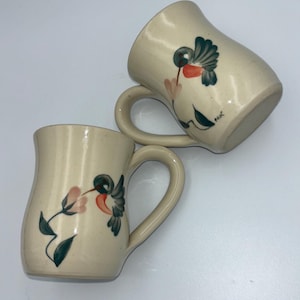 May include: Two ceramic mugs with a cream-colored speckled finish. Each mug features a hand-painted hummingbird design with red, green, and pink accents. The mugs have curved handles and a slightly flared shape.