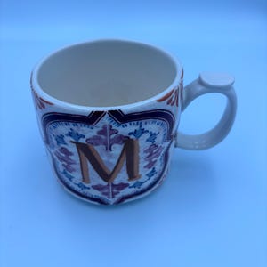 Op de afbeelding: Witte keramische mok met een decoratief ontwerp en de letter "M". De mok heeft een handvat en een gedetailleerd patroon in blauw, paars en oranje. De binnenkant van de mok is wit.