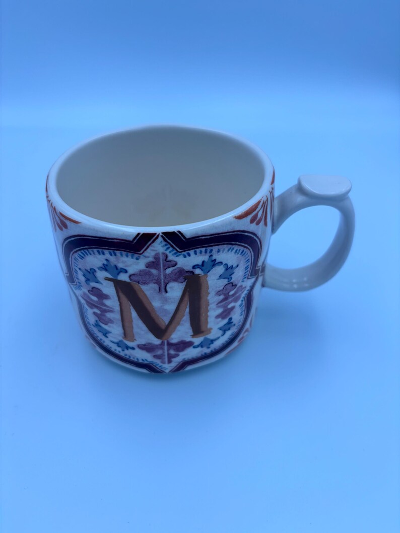 Op de afbeelding: Witte keramische mok met een decoratief ontwerp en de letter "M". De mok heeft een handvat en een gedetailleerd patroon in blauw, paars en oranje. De binnenkant van de mok is wit.