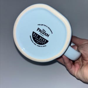 May include: The underside of a light blue ceramic mug featuring black printed text that reads Disney Frozen, Rae Dunn, Artisan Collection by Magenta, 295, may get hot in microwave, and dishwasher safe, made in china, with a white rim.