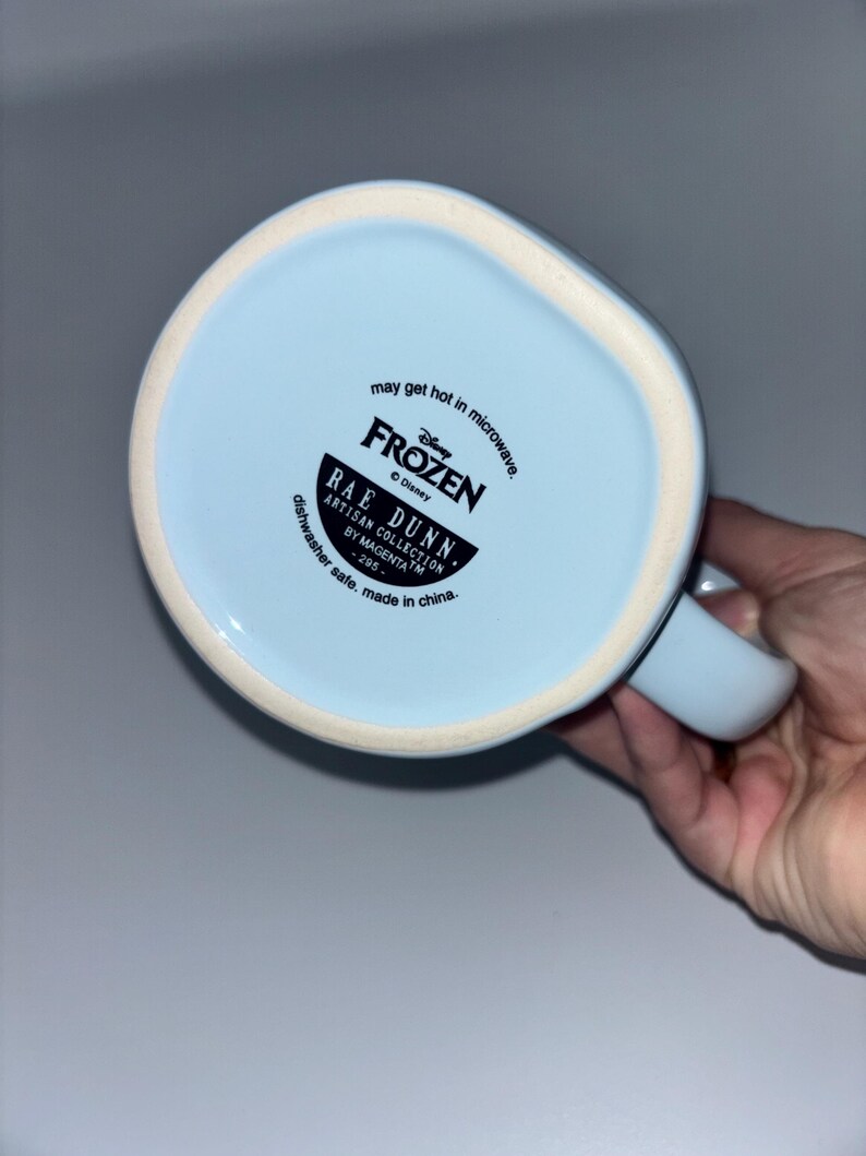 May include: The underside of a light blue ceramic mug featuring black printed text that reads Disney Frozen, Rae Dunn, Artisan Collection by Magenta, 295, may get hot in microwave, and dishwasher safe, made in china, with a white rim.
