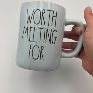 May include: A light blue ceramic mug with a glossy finish and a curved handle. The text "WORTH MELTING FOR" is embossed in black, thin, uppercase lettering on the front of the mug. The design features a simple, cylindrical shape.