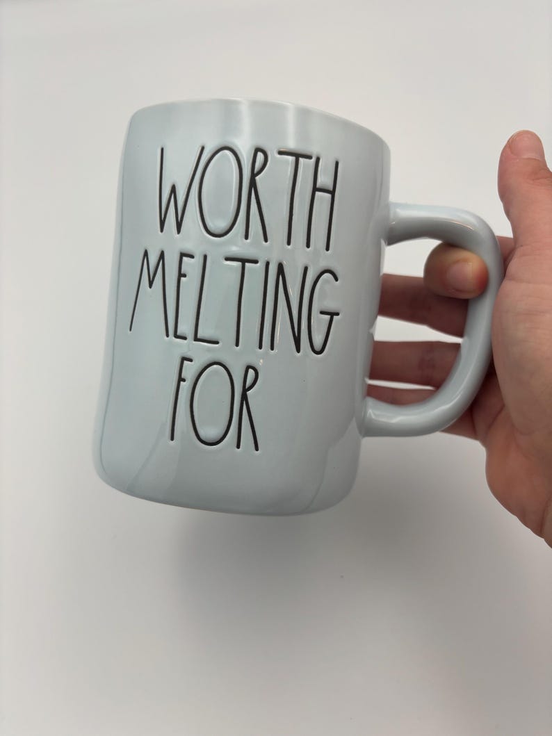 May include: A light blue ceramic mug with a glossy finish and a curved handle. The text "WORTH MELTING FOR" is embossed in black, thin, uppercase lettering on the front of the mug. The design features a simple, cylindrical shape.