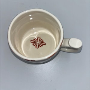 Op de afbeelding: Cr&egrave;mekleurige keramische mok met een handvat. De binnenkant heeft een rood bloemmotief aan de onderkant. De rand heeft een rode accentkleur. De mok is leeg en staat op een lichtgrijze ondergrond.