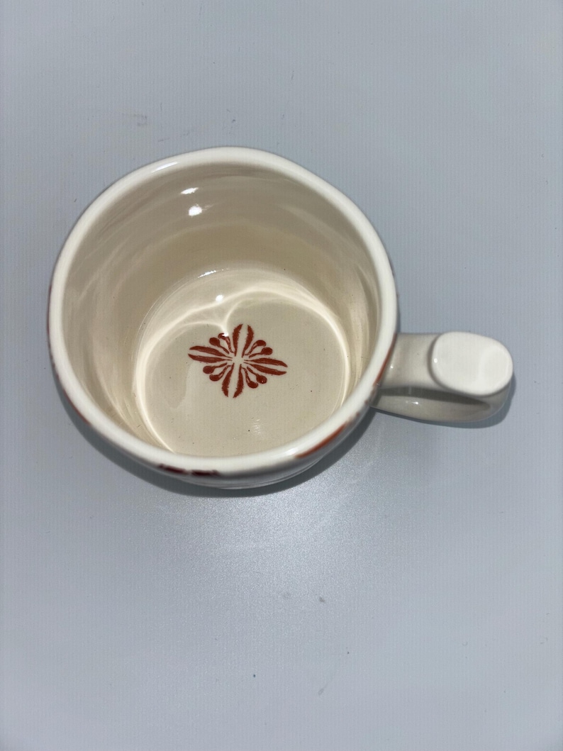 Op de afbeelding: Cr&egrave;mekleurige keramische mok met een handvat. De binnenkant heeft een rood bloemmotief aan de onderkant. De rand heeft een rode accentkleur. De mok is leeg en staat op een lichtgrijze ondergrond.