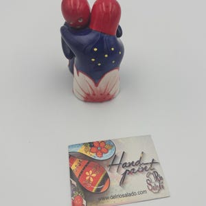 May include: A hand-painted ceramic figurine depicting two figures embracing. The figures have red heads and are dressed in a navy blue outfit with yellow accents. The base is white with a red and white floral design. A card with the text "Hand paint" and "delriosalado.com" is also visible.