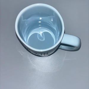 May include: A light blue ceramic mug with a glossy finish, viewed from directly above. The mug features a rounded handle on the right side and a smooth, circular interior. The exterior surface shows a portion of black text printed near the base.