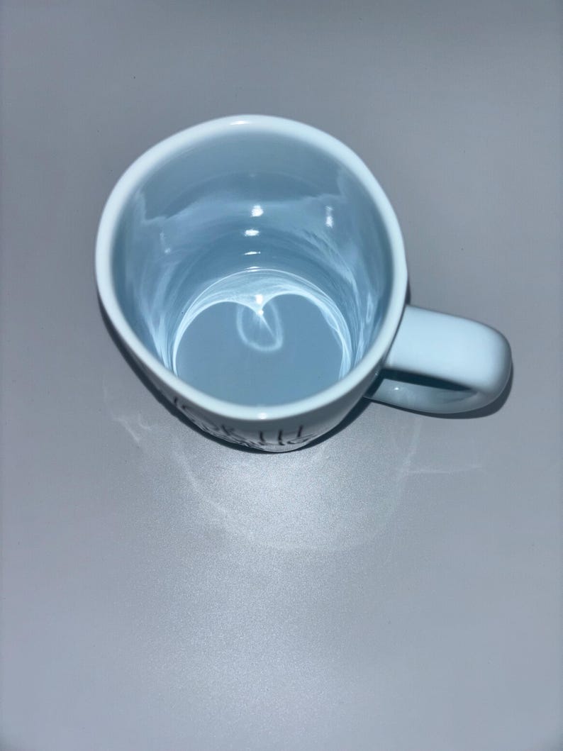 May include: A light blue ceramic mug with a glossy finish, viewed from directly above. The mug features a rounded handle on the right side and a smooth, circular interior. The exterior surface shows a portion of black text printed near the base.