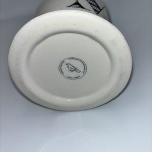 Op de afbeelding: Witte keramische mok met een zwart-wit grafisch ontwerp. De mok heeft een zwart-wit illustratie van een cocktailglas en een gestreept patroon. De bodem van de mok heeft een ronde stempel met een vogelillustratie.