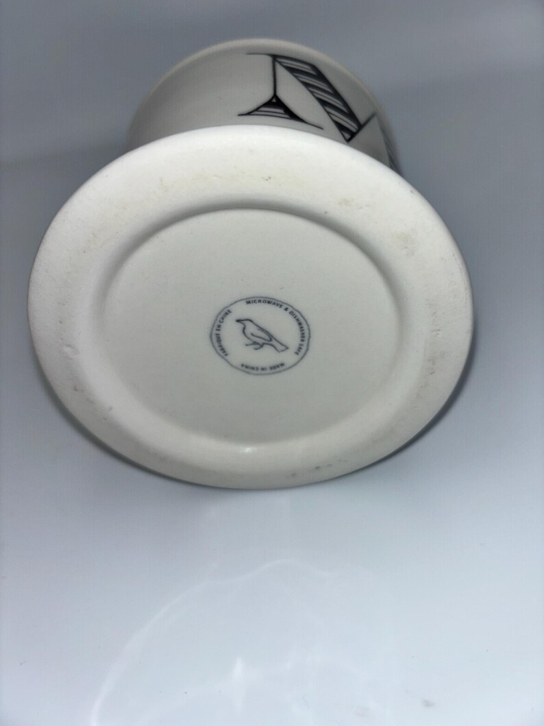 Op de afbeelding: Witte keramische mok met een zwart-wit grafisch ontwerp. De mok heeft een zwart-wit illustratie van een cocktailglas en een gestreept patroon. De bodem van de mok heeft een ronde stempel met een vogelillustratie.