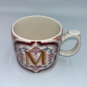Op de afbeelding: Cr&egrave;mekleurige keramische mok met een decoratief ontwerp. De mok heeft een grote gouden letter "M", omgeven door een rood, blauw en wit bloemenpatroon. De handgreep is gebogen met een kleine, platte verlenging.