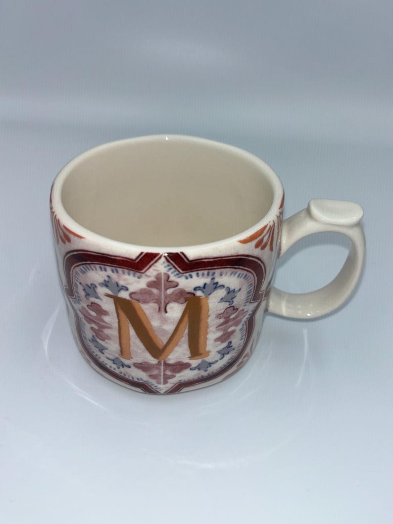 Op de afbeelding: Cr&egrave;mekleurige keramische mok met een decoratief ontwerp. De mok heeft een grote gouden letter "M", omgeven door een rood, blauw en wit bloemenpatroon. De handgreep is gebogen met een kleine, platte verlenging.