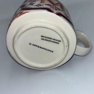 Op de afbeelding: Witte keramische mok met een decoratief patroon in bruin-, rood- en blauwtinten. De mok heeft een oor en de tekst "by ANTHROPOLOGIE" is onderaan zichtbaar. De mok is vaatwasser- en magnetronbestendig.